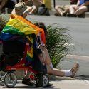 A person sits on a rollator walker beside the road, facing away from the camera to watch the 2022 Halifax Pride parade. They have a rainbow ribbon on their sun hat, and a rainbow flag draped over their shoulders. Hanging on the walker is a tote bag decorated with a the Mi'kmaw eight-pointed star symbol overlaid over birchbark and rainbow blooms, designed by Misiksk Jadis from Abegweit First Nation.