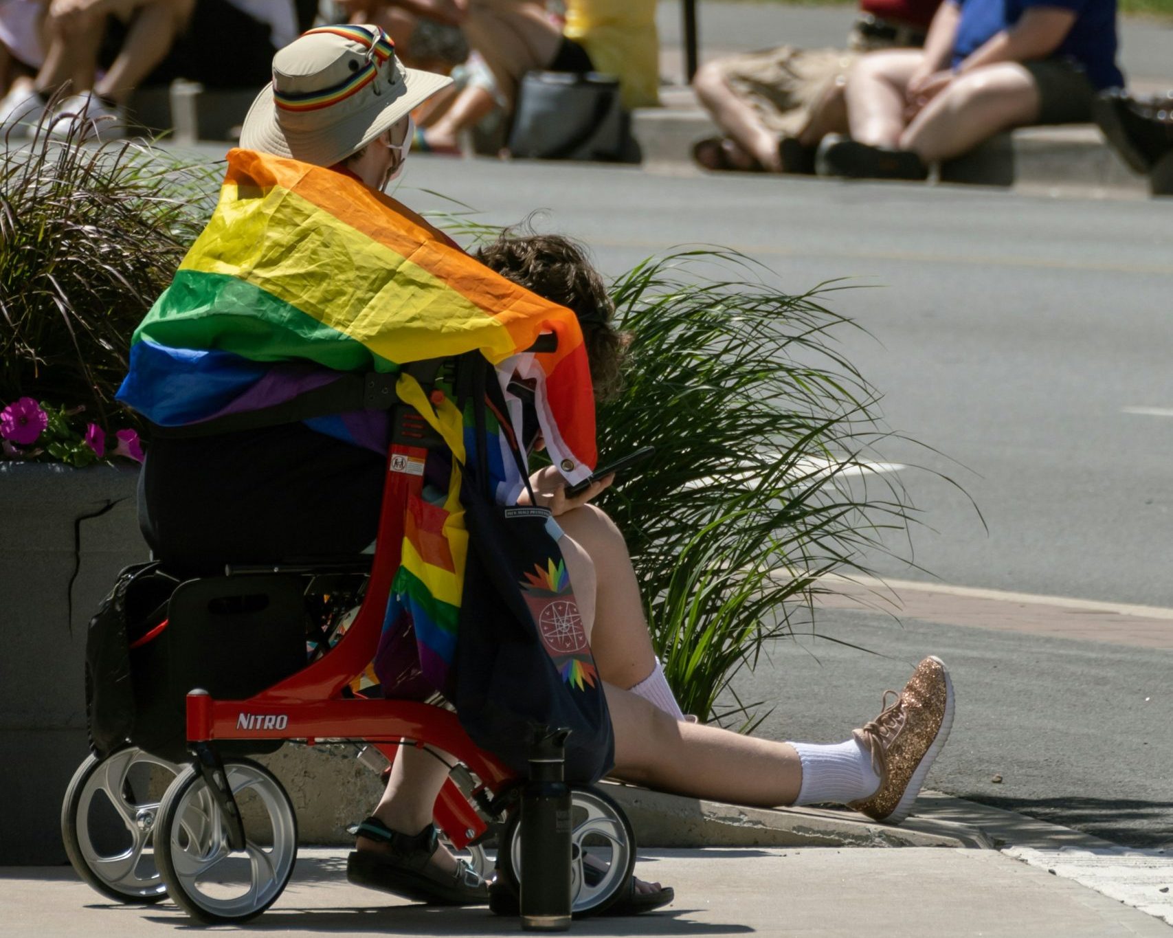 A person sits on a rollator walker beside the road, facing away from the camera to watch the 2022 Halifax Pride parade. They have a rainbow ribbon on their sun hat, and a rainbow flag draped over their shoulders. Hanging on the walker is a tote bag decorated with a the Mi'kmaw eight-pointed star symbol overlaid over birchbark and rainbow blooms, designed by Misiksk Jadis from Abegweit First Nation.